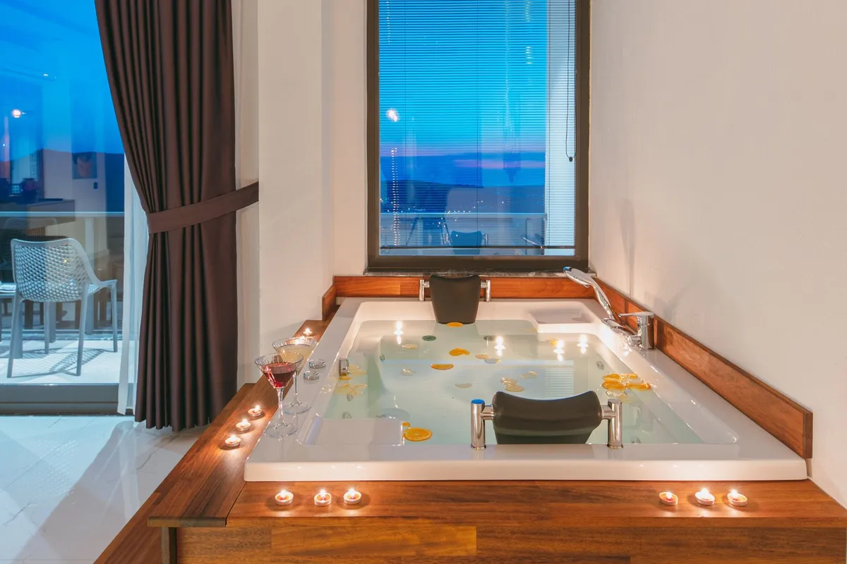 A private in-room jacuzzi suite dressed for a honeymoon at dusk, with tealight candles along the wooden rim, two drinks poured and citrus slices floating in the water